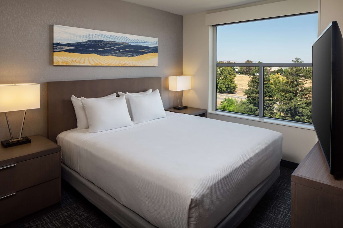 Een hotelkamer met een groot bed, twee nachtkastjes met lampen, een flatscreen televisie en een groot raam met uitzicht op de omgeving.