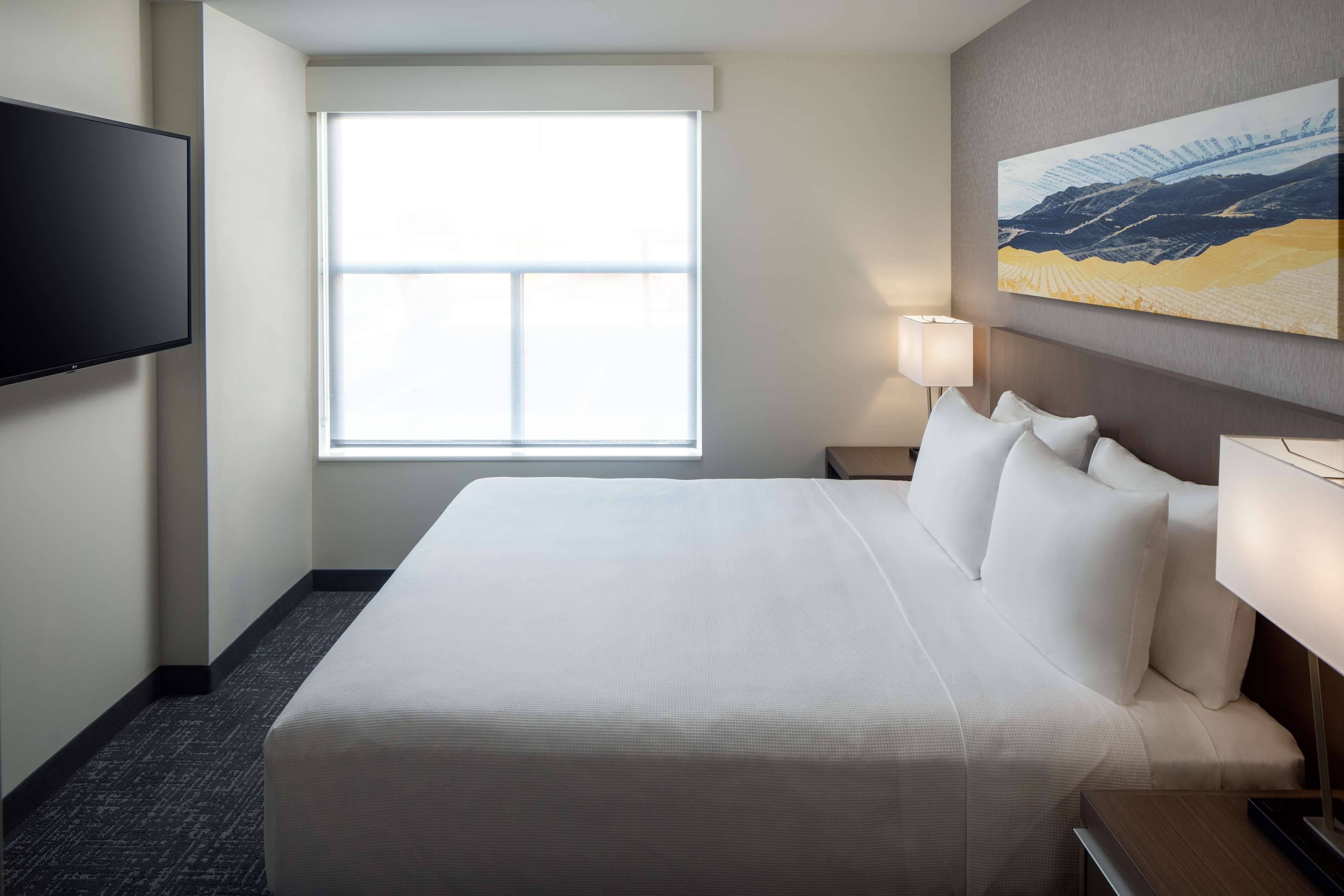 Habitación de hotel con una cama grande, un televisor de pantalla plana montado en la pared, una ventana con persianas, una lámpara de noche y un cuadro en la pared.