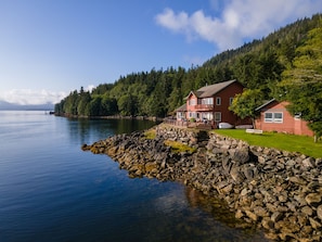 A coastal house with a stone dock, a red building, and a forested hillside. - Salmon Run Vacation Rental! (Ketchikan)