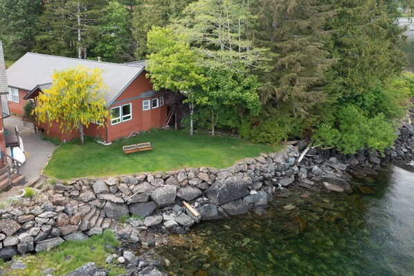A cabin with a stone wall and a grassy yard, surrounded by trees and a body of water. - Salmon Run Vacation Rental! (Ketchikan)