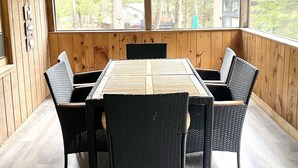 Ein Zimmer mit Holzwandverkleidung, einem großen Tisch, vier Stühlen und einem Pendel über dem Tisch. Große Fenster bieten Blick auf einen Wald.