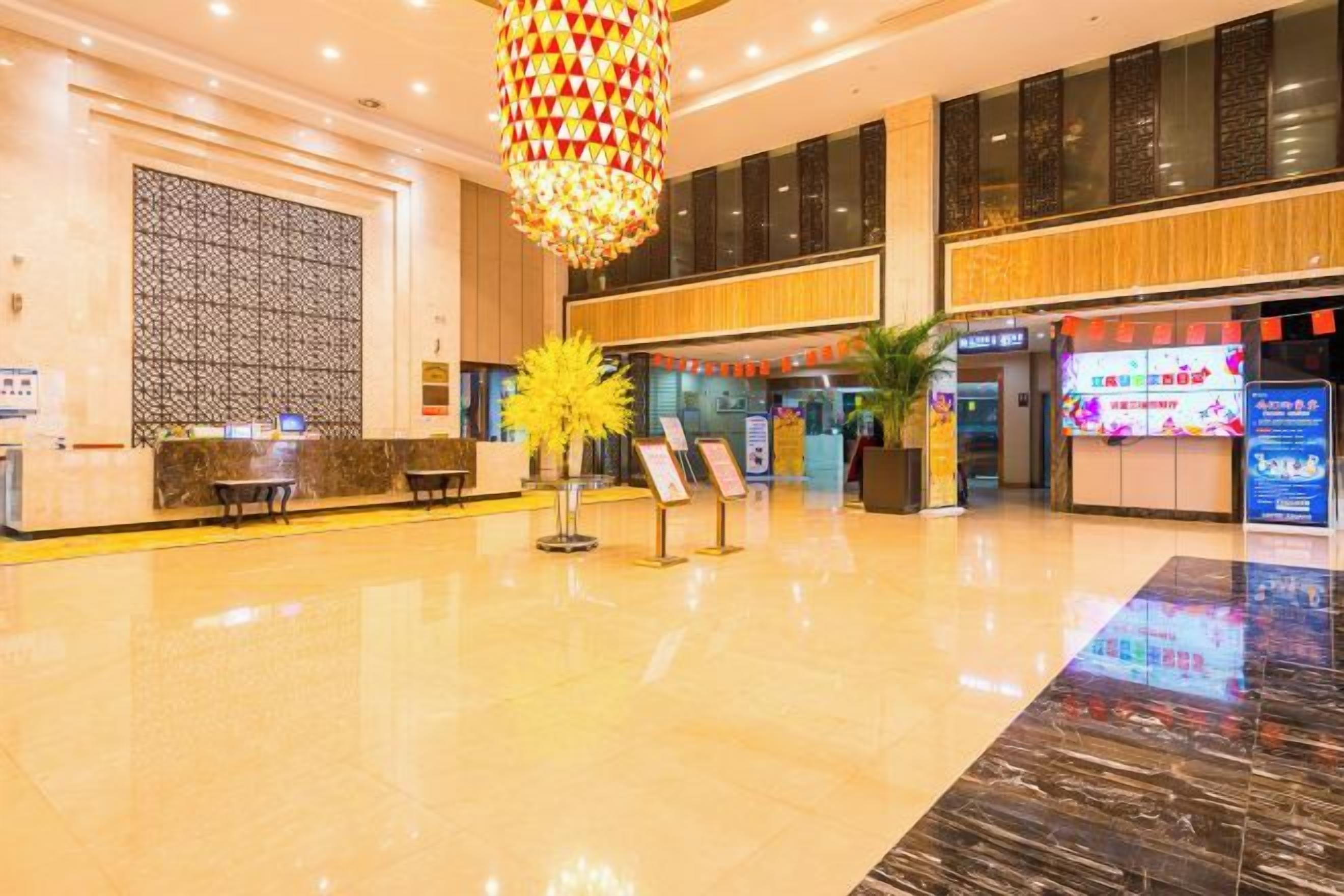 A hotel lobby with a reception desk, a large chandelier, and a digital information board.