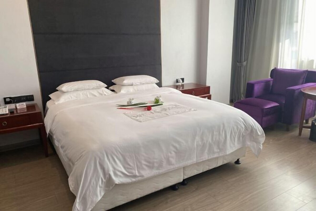 Une chambre d’hôtel avec un grand lit, une table de chevet, un bureau, une chaise violette et une fenêtre avec des rideaux.