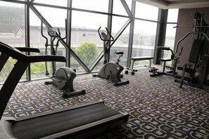 Ein Fitnessstudio mit Laufband, Crosstrainer und Krafttrainingsgeräten, großen Fenstern und einem gemusterten Boden.