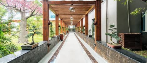A covered walkway with a stone wall on one side and a building on the other, leading to a garden area with trees and plants.