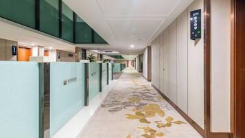 Un corridoio di un hotel con pavimento rivestito di moquette, pareti divisorie in vetro e una segnaletica che indica la direzione dell'ascensore.