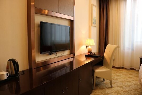 Une chambre d’hôtel équipée d’un téléviseur à écran plat fixé à un meuble en bois, d’un bureau avec une lampe, d’une chaise et d’un lit recouvert d’une moquette à motifs.