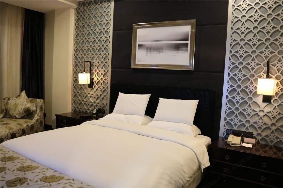 Una habitación de hotel con una cama grande, cabecera, dos mesitas de noche con lámparas, una silla y un panel decorativo en la pared.
