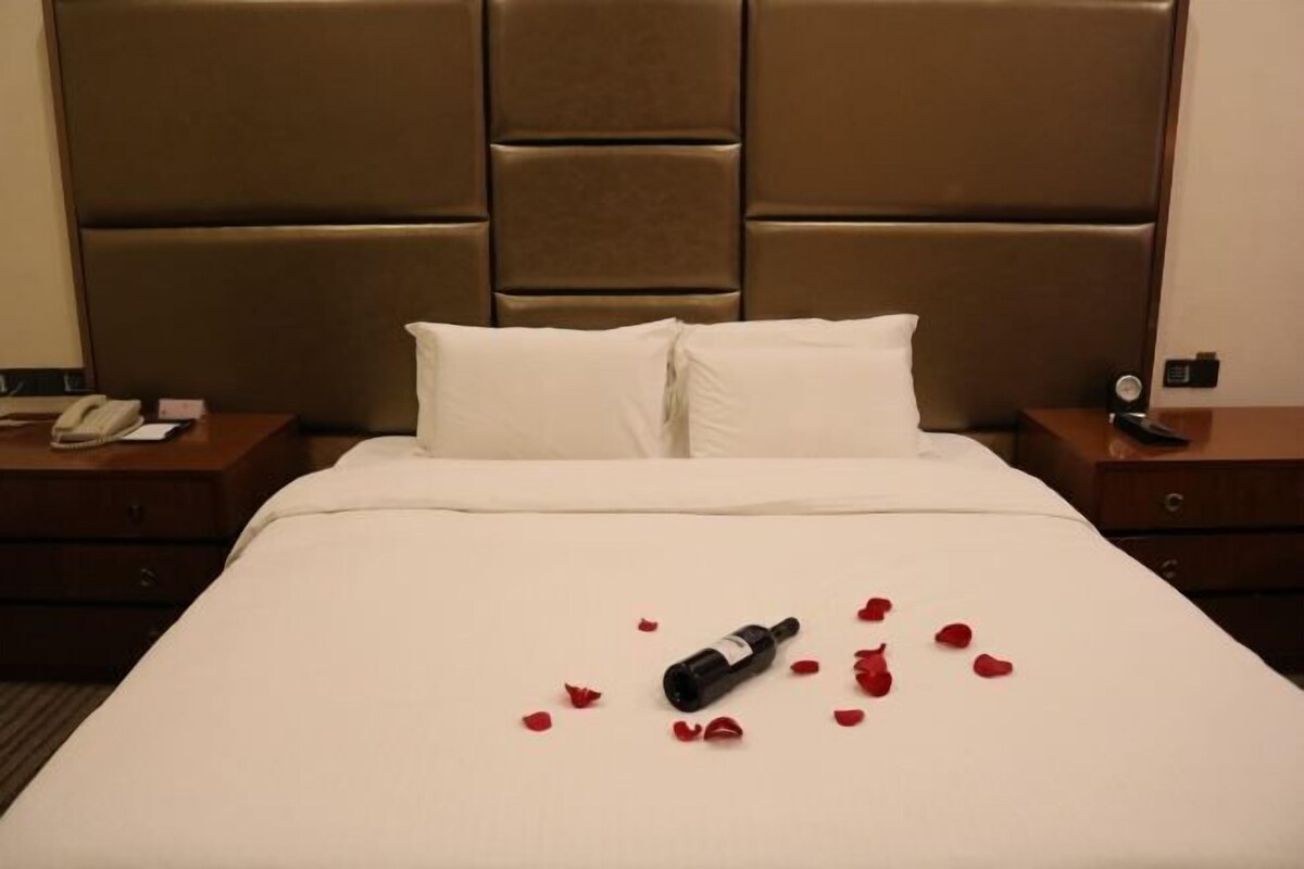 Une chambre d’hôtel avec un grand lit, deux oreillers, une table de chevet avec un téléphone et une bouteille de vin, le tout agrémenté de pétales de rose disposés sur le lit.