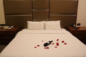 Une chambre d’hôtel avec un grand lit, deux oreillers, une table de chevet avec un téléphone et une bouteille de vin, le tout agrémenté de pétales de rose disposés sur le lit.
