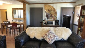 A cozy living room with a black leather couch, a wooden dining table with chairs, a microwave, and a decorative fan.