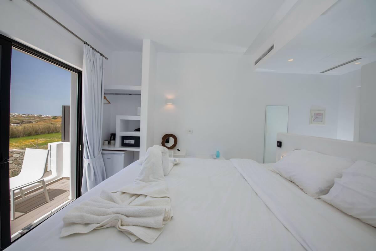 A modern hotel room with a large bed, a small desk, a mirror, and a view of the outdoors.