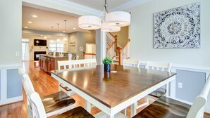 Un espace repas comprenant une grande table en bois, des chaises blanches, un éclairage suspendu et un élément décoratif mural.