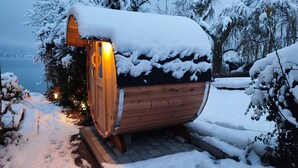 A wooden cabin with a snow-covered roof and a lit interior, surrounded by snow-laden trees and a view of a body of water.