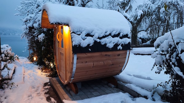 A wooden cabin with a snow-covered roof and a lit interior, surrounded by snow-laden trees and a view of a body of water.