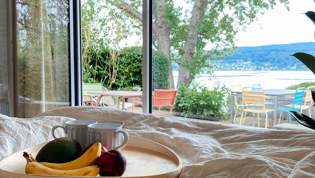 A bedroom with a large window overlooking a garden and a lake. There is a wooden tray with fruits and a mug on the bed.