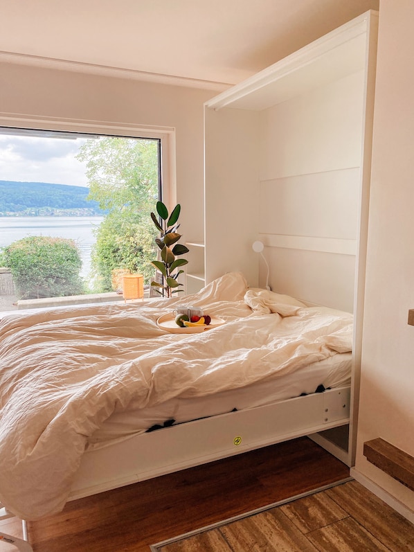 Un lit avec un cadre en bois, une table de chevet ornée d’une plante et une fenêtre donnant sur un lac et des collines.