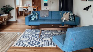 A modern living room with a blue sofa, wooden flooring, and a dining area in the background.
