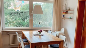 A dining area with a wooden table, four chairs, a hanging lamp, and a view of greenery outside.