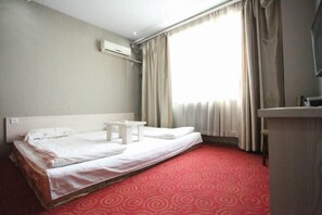 Habitació d'hotel amb un llit individual, una petita taula de nit, un aparell d'aire condicionat a la paret, un escriptori amb una cadira i un gran finestral amb cortines.