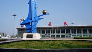 Una escultura azul abstracta, un edificio con fachada de vidrio y un cielo despejado.