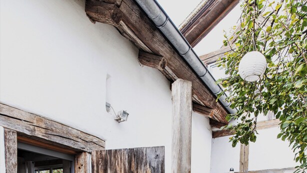 Una puerta de madera rústica da a un espacio con piso de baldosas, un pequeño jardín con plantas y un banco, y un farol colgante.