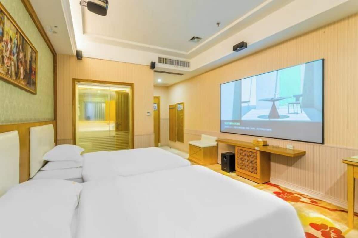 A hotel room with a large bed, a television screen, a wooden desk, and a door leading to another room.