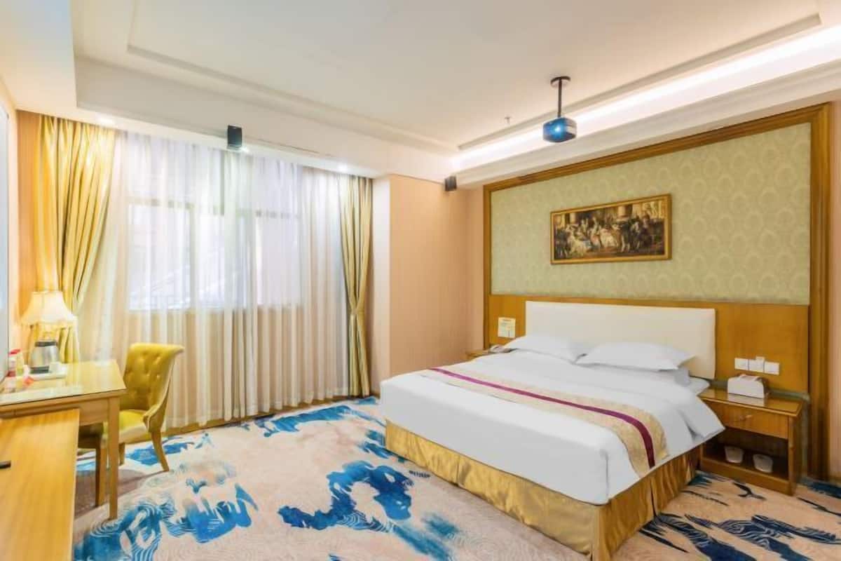 A hotel room with a bed, a desk, a chair, a nightstand, and a painting on the wall.