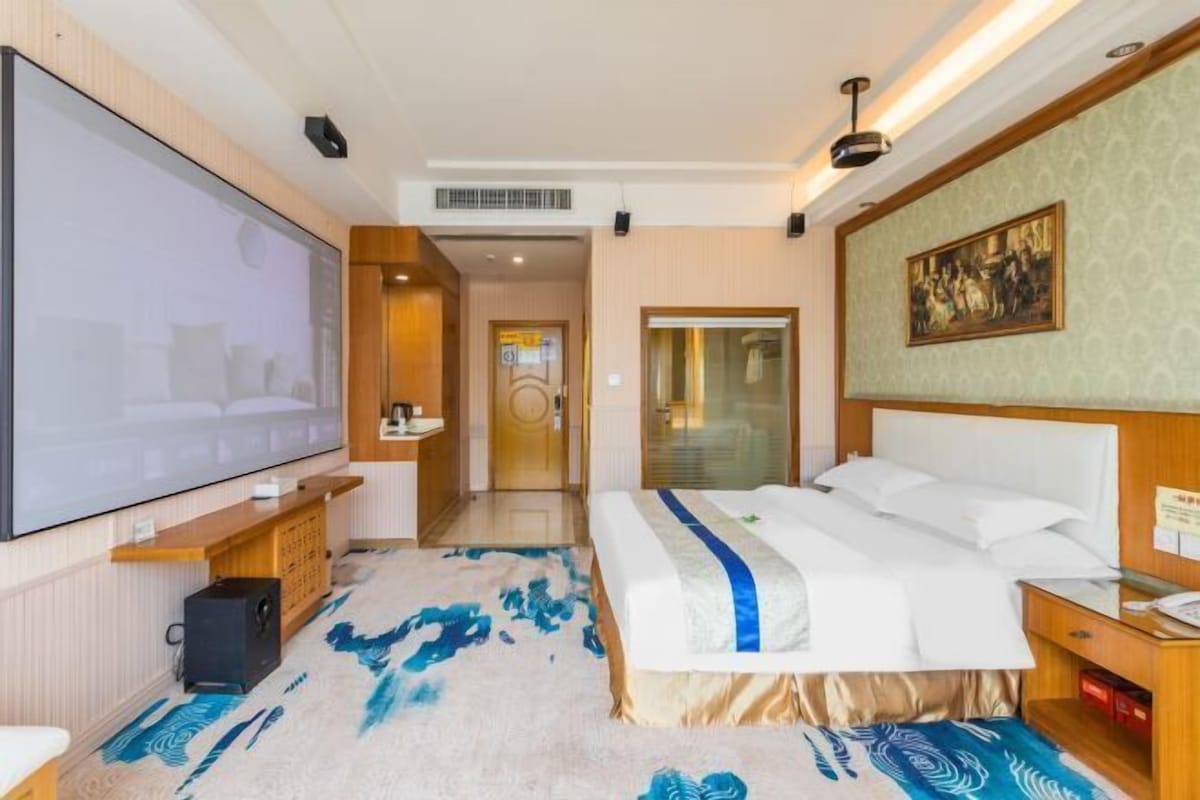 A hotel room with a large flat-screen TV, a bed with a blue and white bedspread, a wooden nightstand, a desk with a lamp, and a painting on the wall.