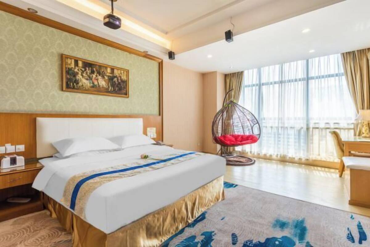 A hotel room with a large bed, a hanging chair, a painting on the wall, and a view of the city through large windows.