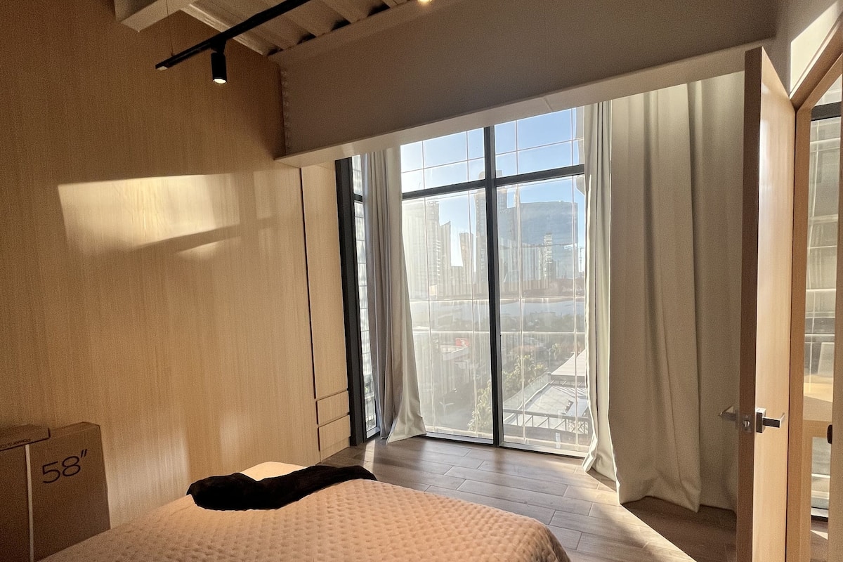 A bedroom with a large bed, a window with curtains, and a view of the city.