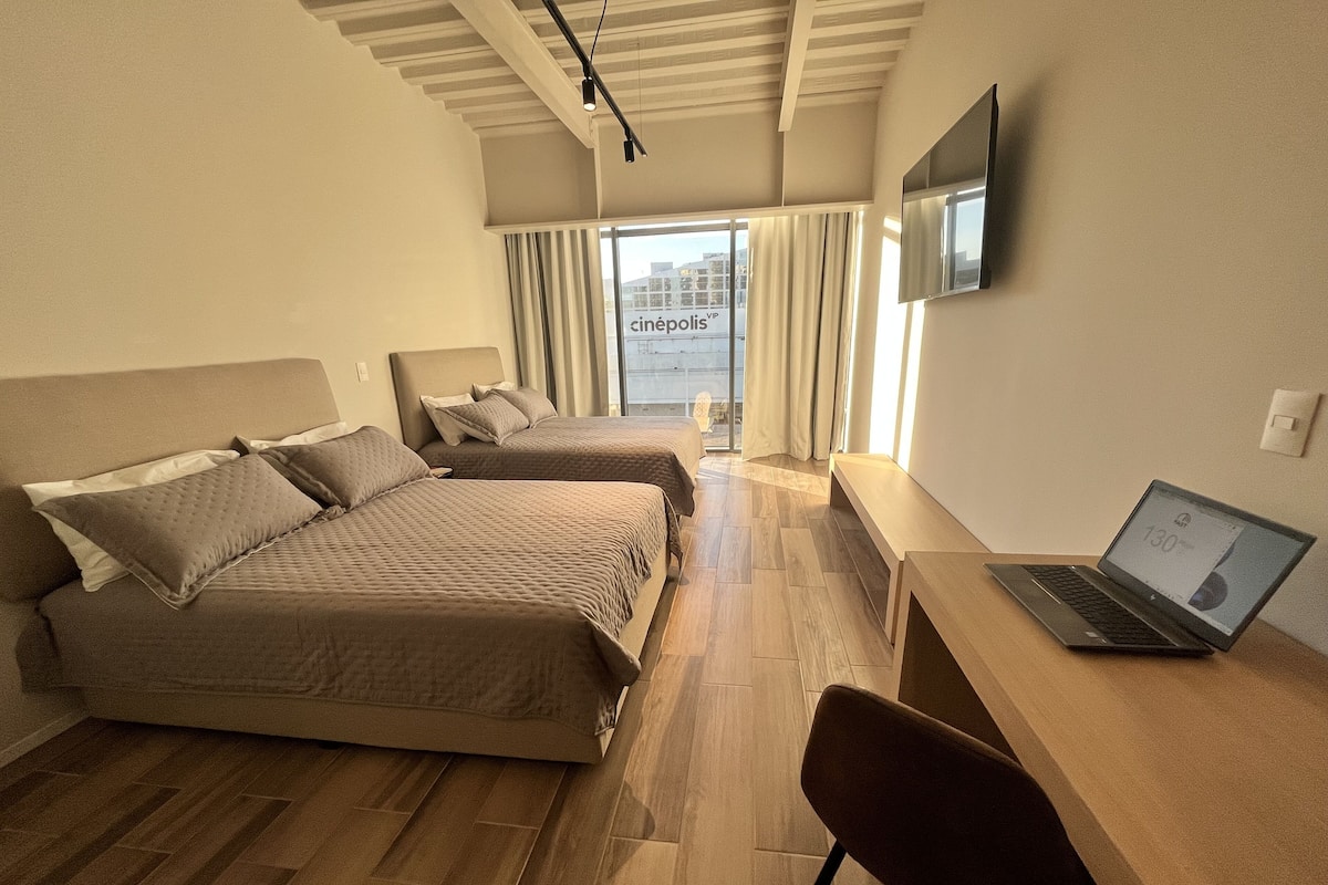 A hotel room with two beds, a desk with a laptop, a TV on the wall, and a view of the city through the window.