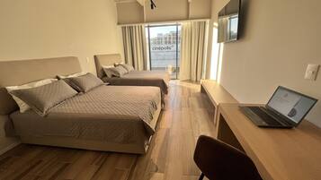 Quarto de hotel com duas camas, uma escrivaninha com um laptop, uma televisão na parede e vista para a cidade pela janela.
