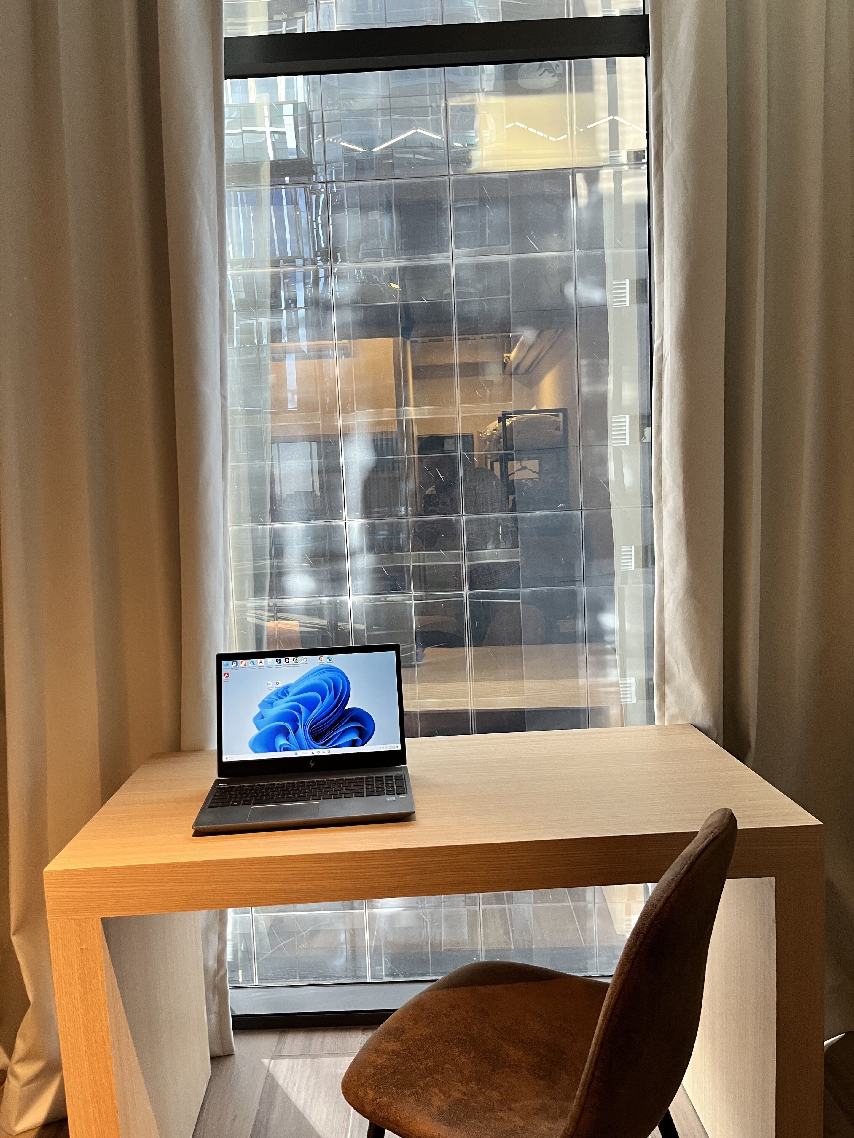 Een laptop op een houten bureau met een stoel ervoor, geplaatst bij een raam met uitzicht op een gebouw.