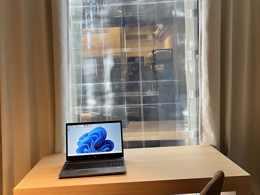 Een laptop op een houten bureau met een stoel ervoor, geplaatst bij een raam met uitzicht op een gebouw.