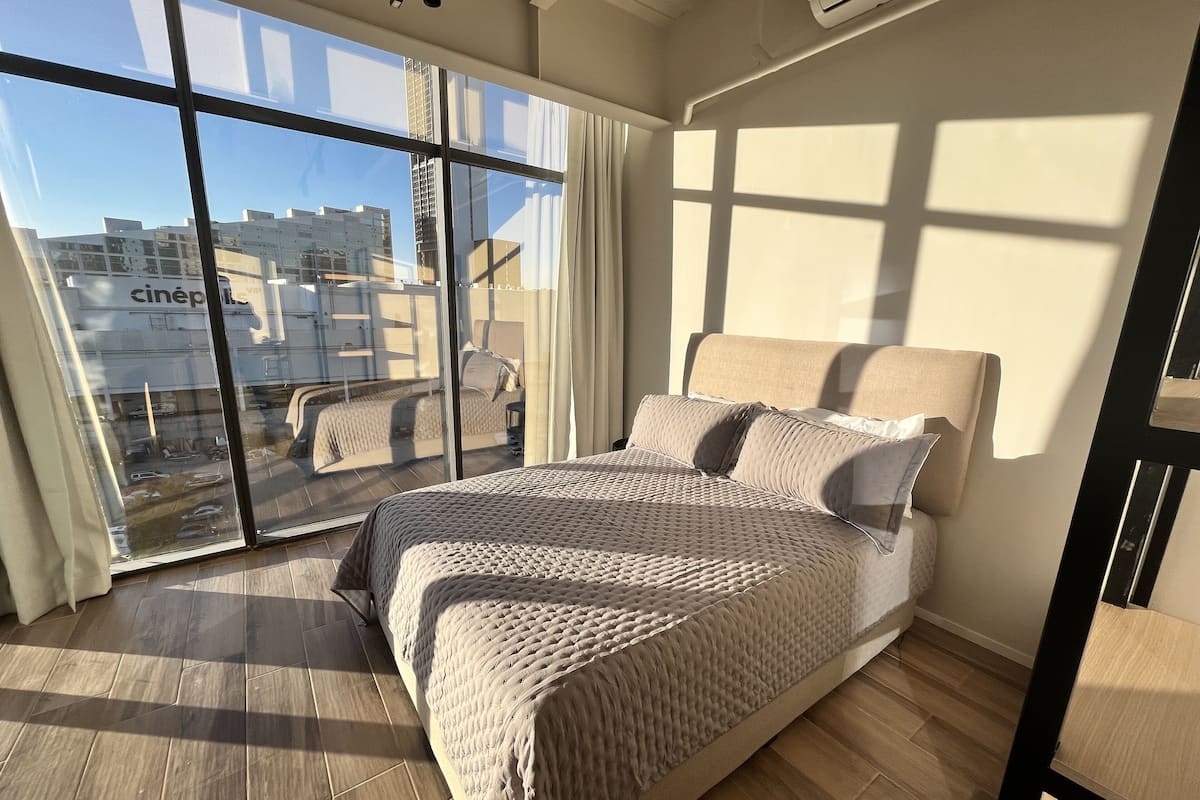 A bedroom with a large window, a bed with a textured blanket, and a view of buildings outside.