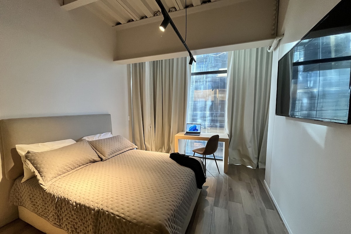 A modern hotel room with a large bed, a desk with a laptop, and a view of the city through large windows.