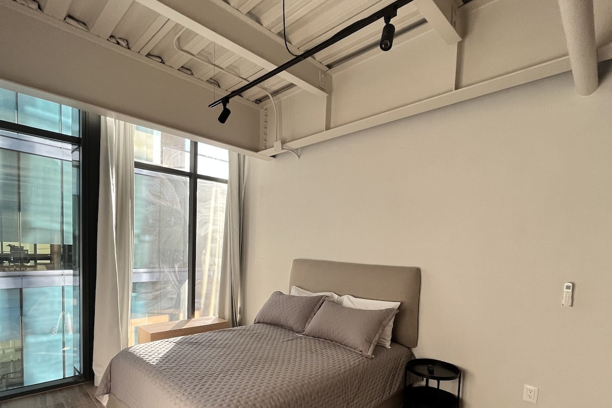 A modern bedroom with a large bed, a view through the window, and a ceiling with exposed beams.