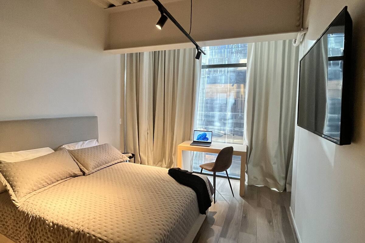 A modern hotel room with a large bed, a desk with a laptop, and a TV on the wall.