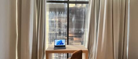 A modern hotel room with a bed, a desk with a laptop, and a view of the city through large windows.
