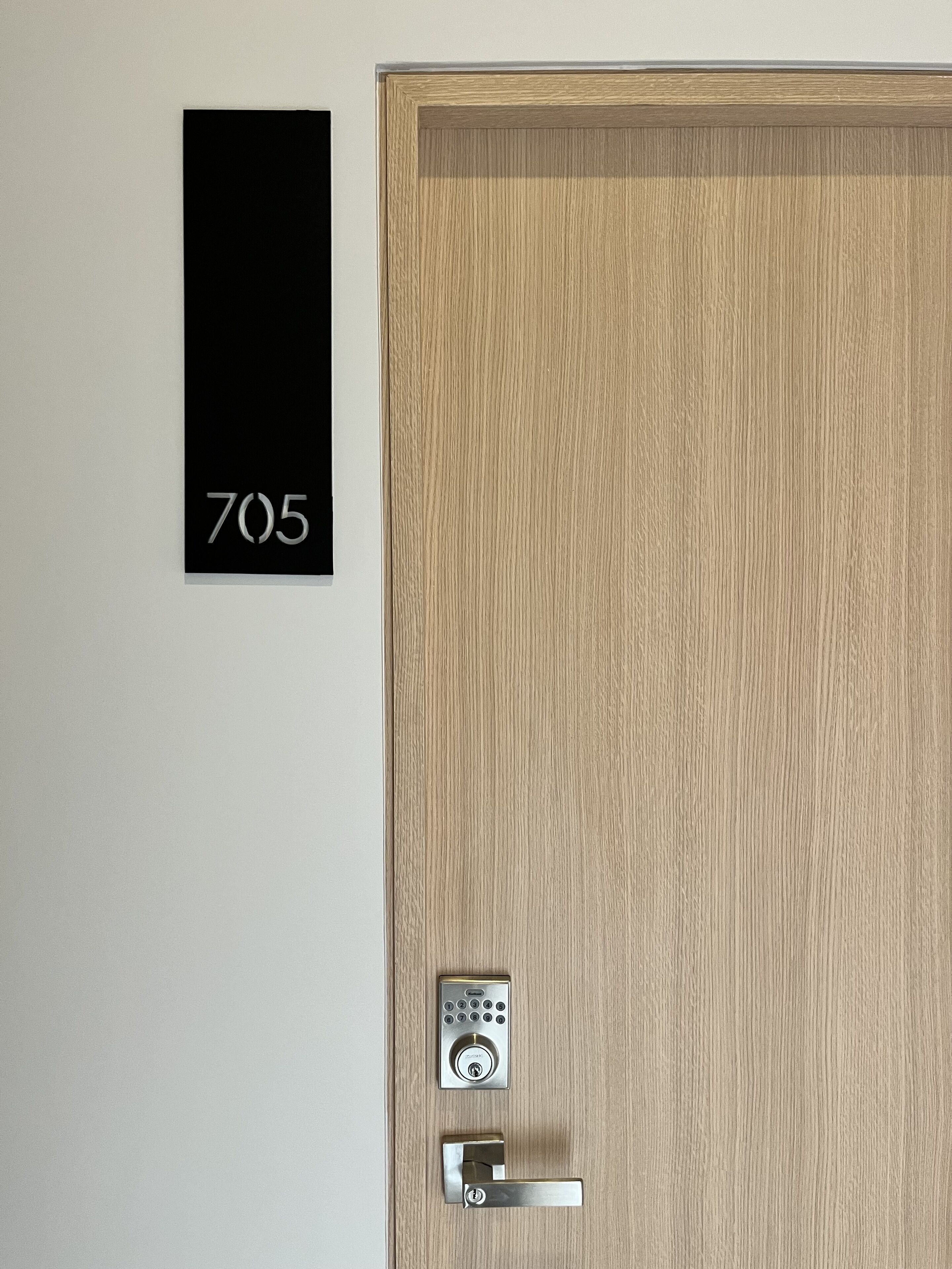 Une porte en bois dotée d’un clavier numérique et d’un trou de serrure. Une plaque noire portant le numéro 705 est fixée au mur, sur le côté gauche.
