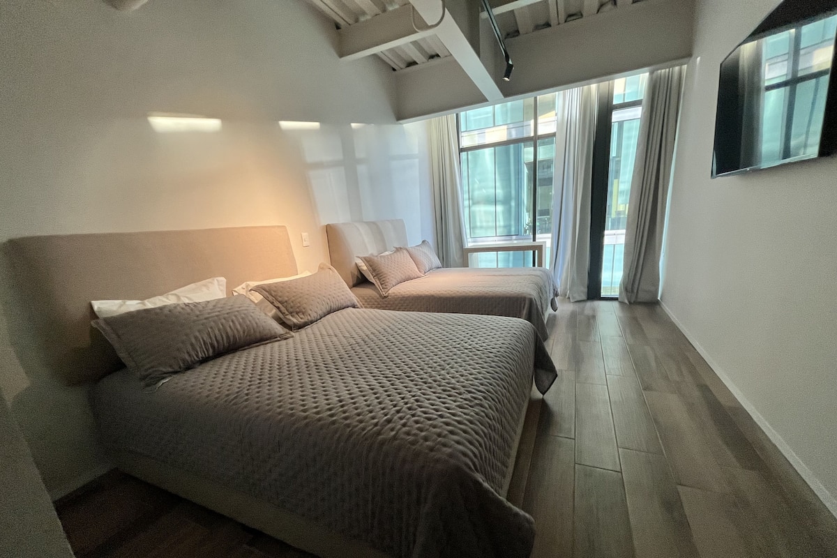 A modern hotel room with two beds, a large window with curtains, and a flat-screen TV mounted on the wall.