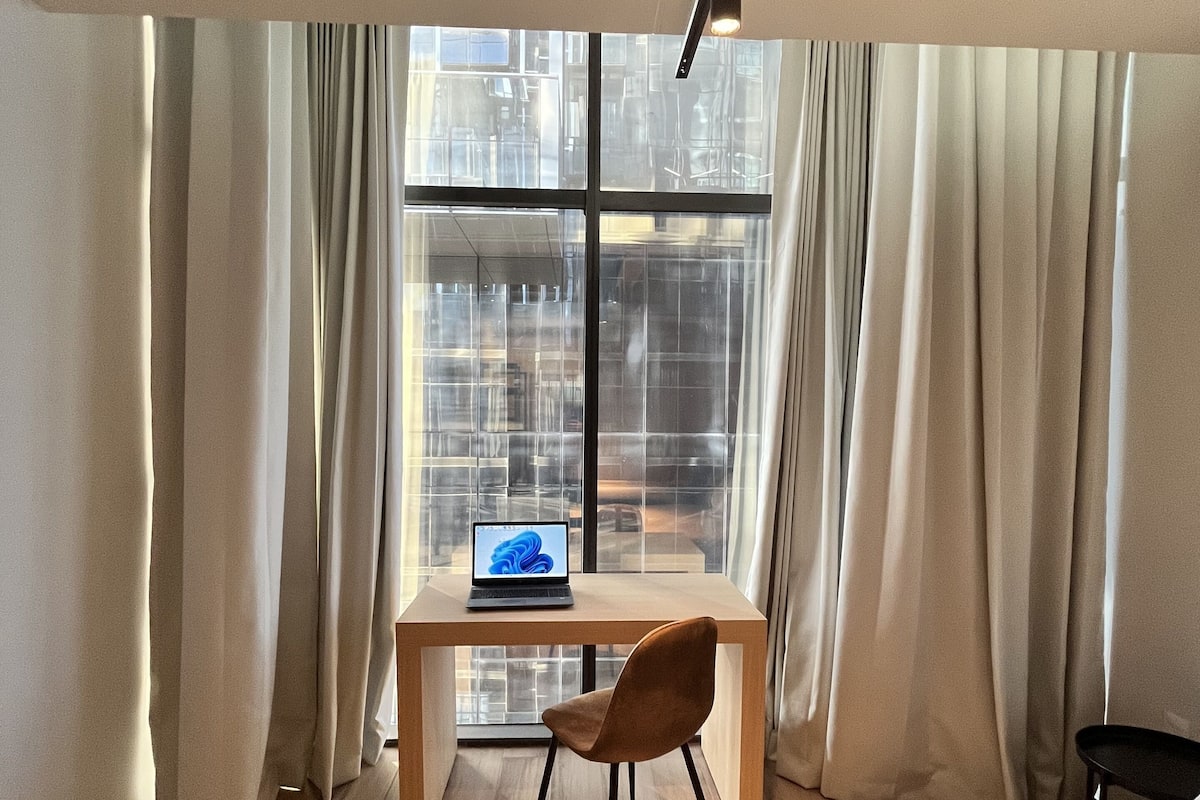 A hotel room with a bed, a desk with a laptop, a chair, and a large window with curtains.