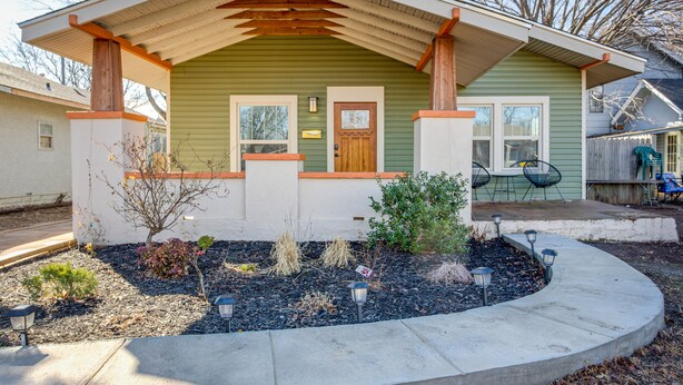 A single-story house with a green exterior and a wooden porch. There is a small garden with various plants and a pathway leading to the front door. - Chic Lawton Home, 5 Mins to Fort Sill! (Lawton)