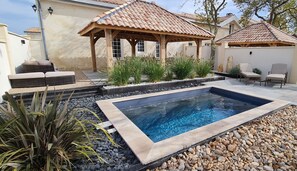 A swimming pool with a wooden deck, surrounded by rocks and plants, and a covered seating area with chairs.
