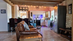 A living room with a leather sofa, a dining area with a table and chairs, a kitchen with a washing machine, and a wooden ceiling.