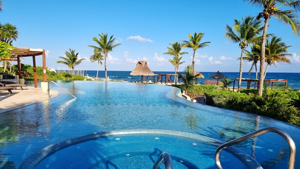 A clear blue infinity pool with a view of the ocean, surrounded by palm trees and thatched-roof structures.