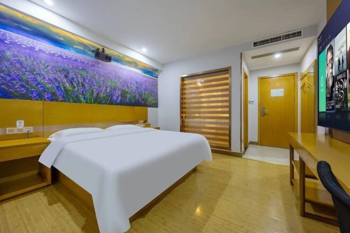 A hotel room with a bed, a desk, a chair, a large window with blinds, and a wall mural.