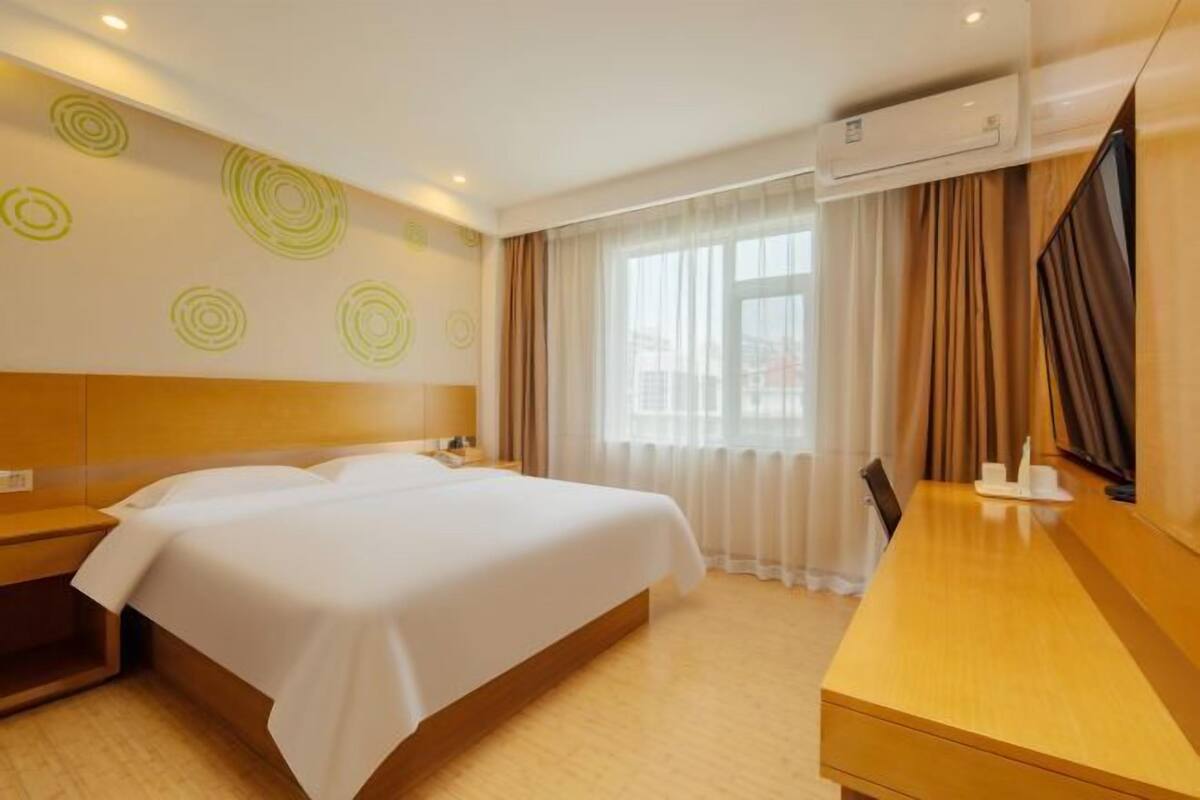 A hotel room with a bed, bedside tables, a desk, an air conditioning unit, and a window with curtains.