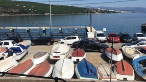 Un puerto deportivo con varias embarcaciones amarradas, un estacionamiento con coches, y una vista despejada del agua y colinas lejanas.